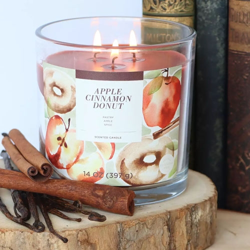 A lit scented candle labeled \“Apple Cinnamon Donut\“ placed on a wooden surface next to cinnamon sticks and vintage books.