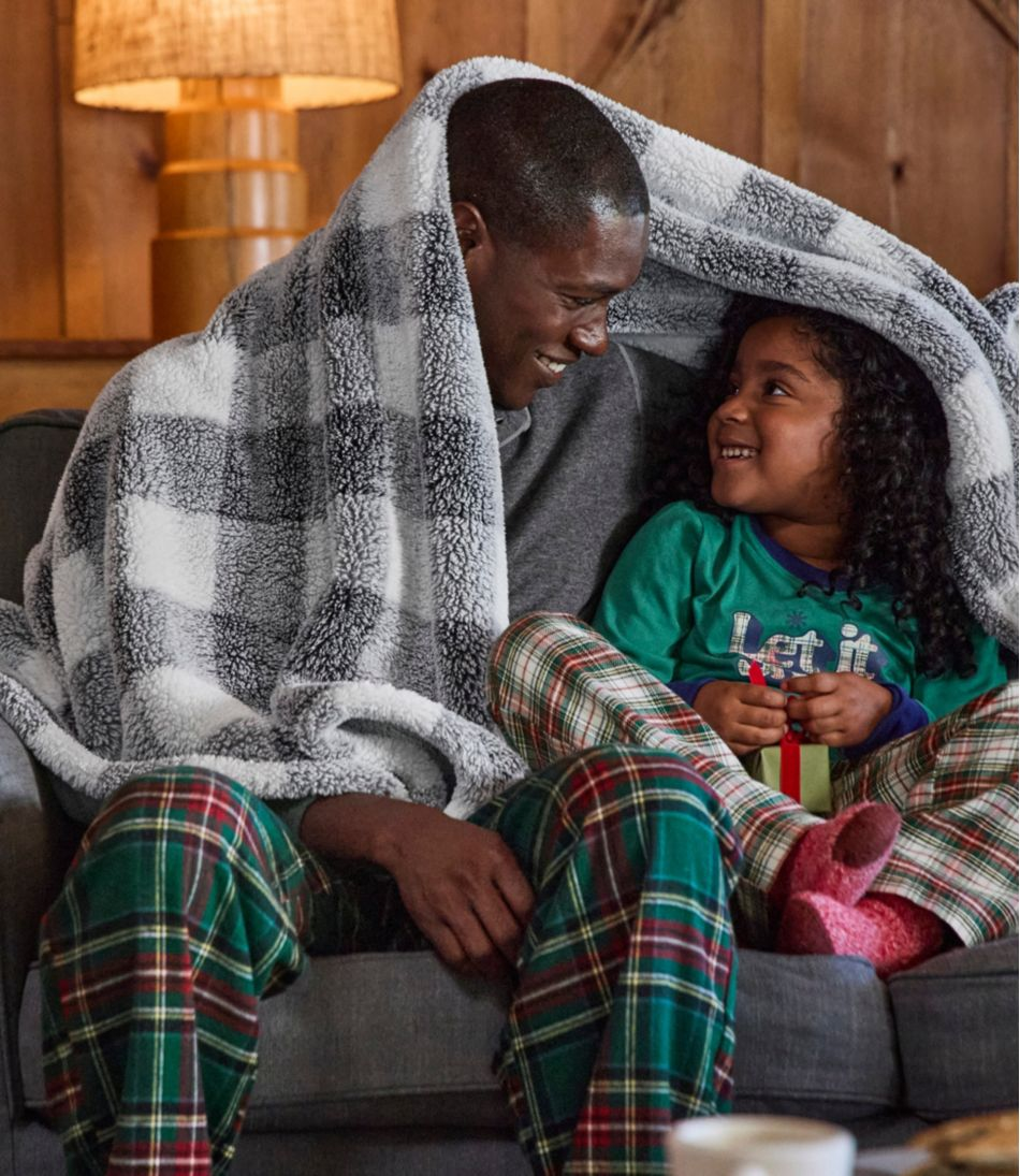 Two people are sitting on a couch, wrapped in a black and white plaid blanket, both wearing plaid pajamas.