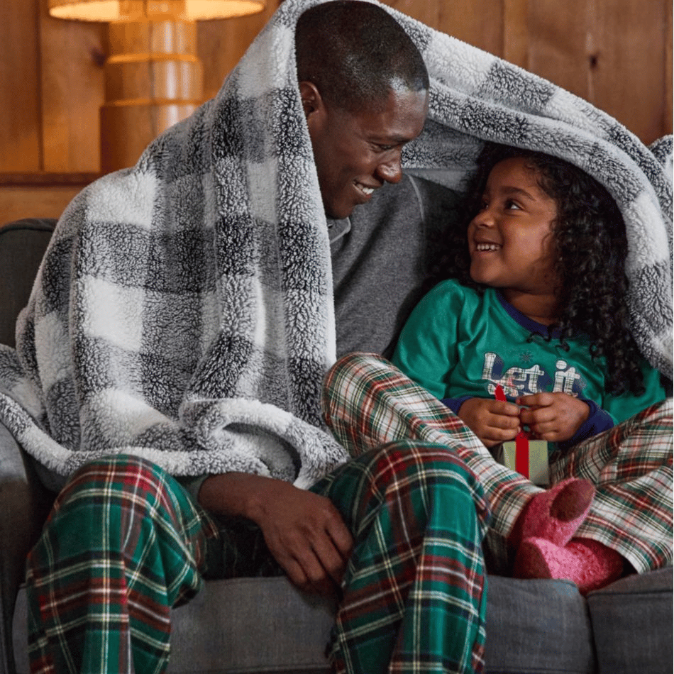 Two people are sitting on a couch, wrapped in a black and white plaid blanket, both wearing plaid pajamas.