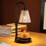 A black lamp with a textured glass shade illuminates a lit candle placed on its base. There are magazines stacked in the background.