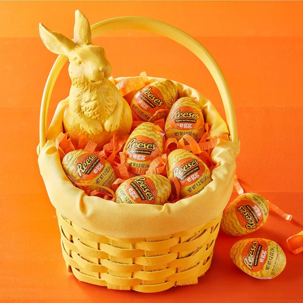 A yellow basket contains Reese's eggs and a chocolate bunny, all wrapped in orange foil, with some eggs scattered around it.