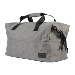A gray, spacious, rectangular duffel bag with black handles and an adjustable shoulder strap. It features a front patch and a modern, minimalist design.