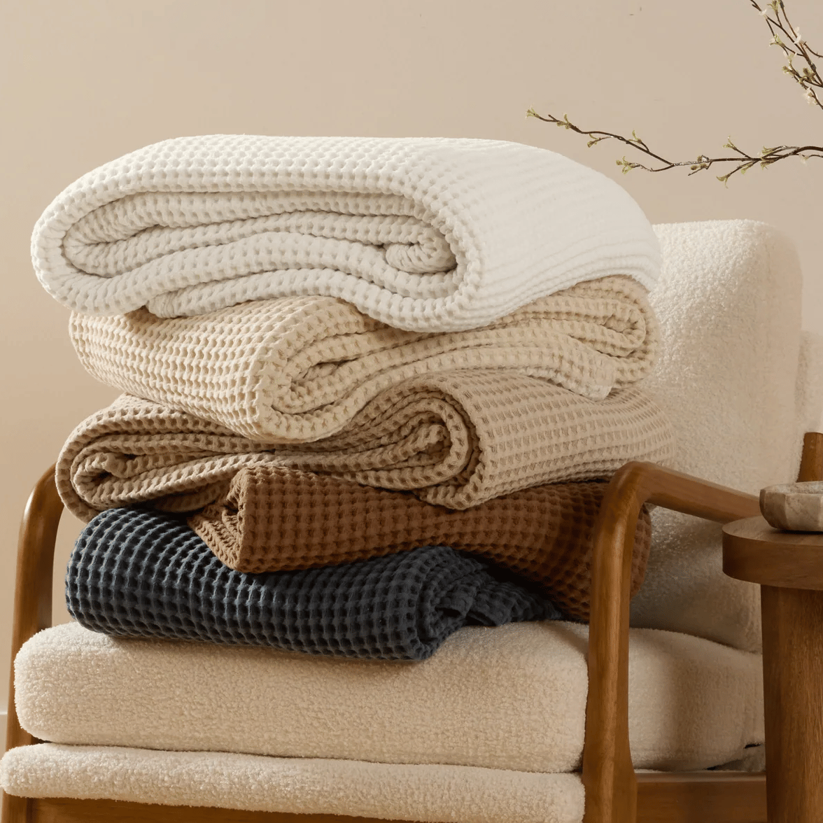Five folded waffle-knit blankets in white, cream, beige, brown, and dark blue are stacked on a cushioned chair with wooden arms.