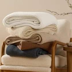 Five folded waffle-knit blankets in white, cream, beige, brown, and dark blue are stacked on a cushioned chair with wooden arms.