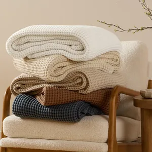 Five folded waffle-knit blankets in white, cream, beige, brown, and dark blue are stacked on a cushioned chair with wooden arms.