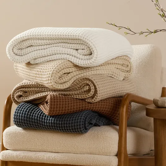 Five folded waffle-knit blankets in white, cream, beige, brown, and dark blue are stacked on a cushioned chair with wooden arms.