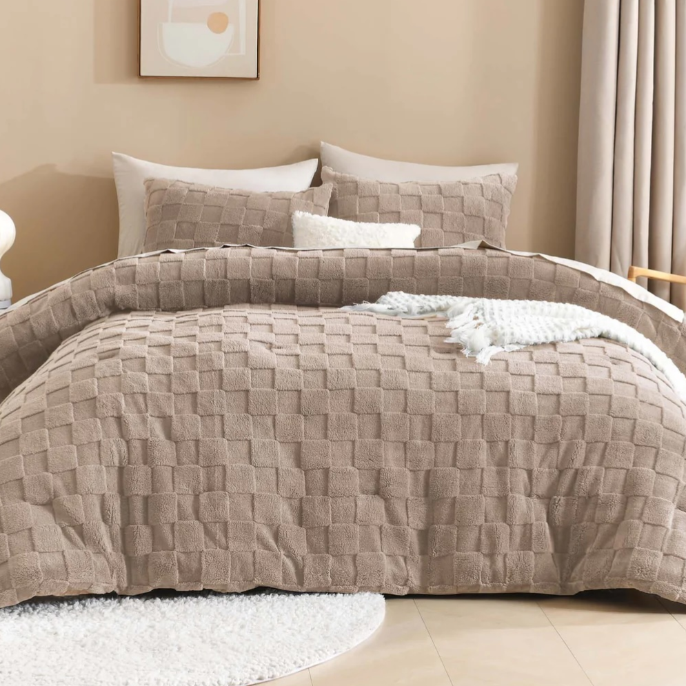 A beige quilted bedding set with matching pillows, a cozy white textured throw, and a minimalistic art piece on the wall, accompanied by a small side table with a lamp. Neutral curtains and a round rug complete the cozy bedroom decor.