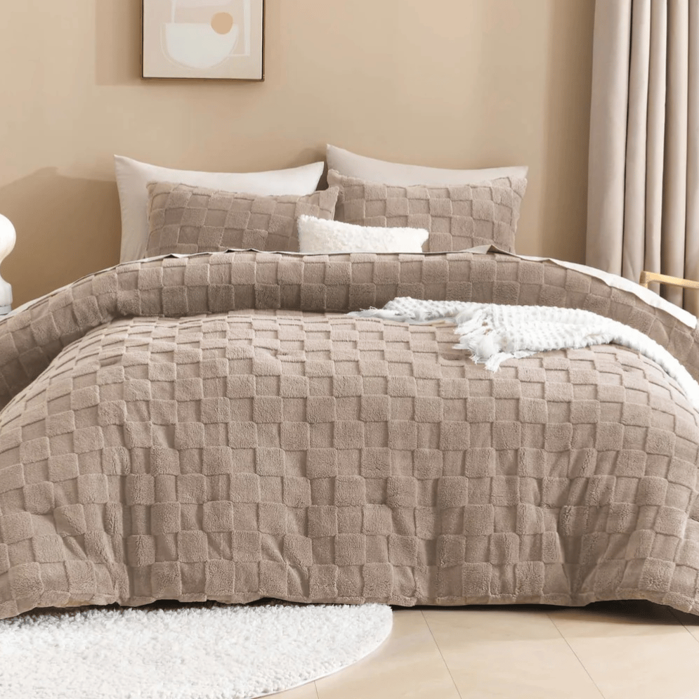 A beige quilted bedding set with matching pillows, a cozy white textured throw, and a minimalistic art piece on the wall, accompanied by a small side table with a lamp. Neutral curtains and a round rug complete the cozy bedroom decor.