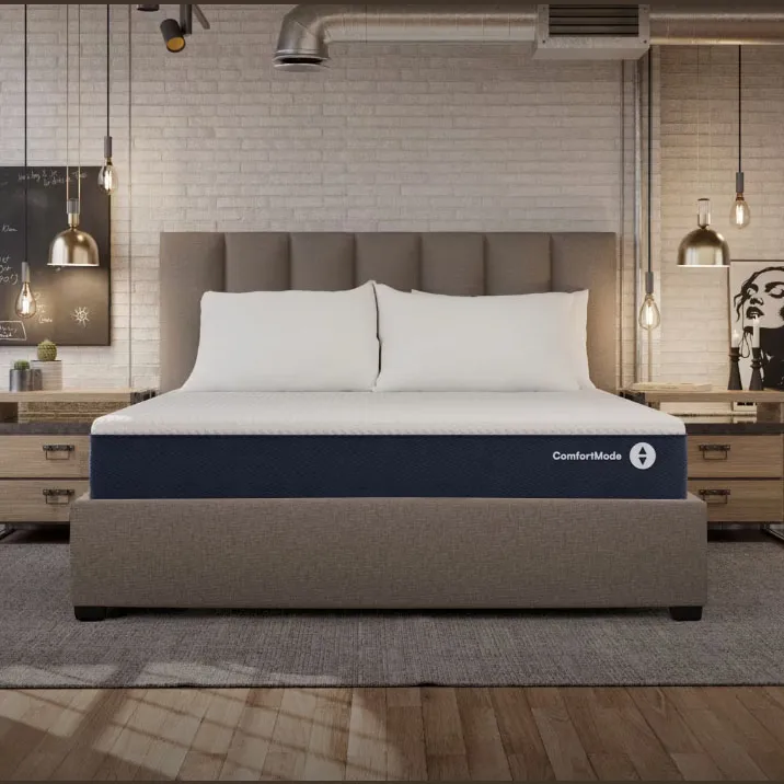 A contemporary bedroom features a king-sized bed with a ComfortMode mattress, upholstered headboard, and two side tables with industrial-style pendant lights. A vent and exposed brick wall add an industrial touch, alongside minimalist decor.