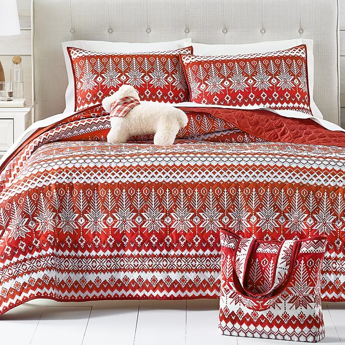 A queen-size quilt set with a festive red and white snowflake pattern, including two matching pillow shams and a tote bag, accessorized with a small white plush dog.