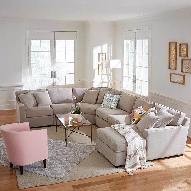 A beige 5-piece Radley sectional sofa features a modern L-shaped design with plush cushions and various colorful throw pillows, complemented by a clear glass coffee table. A small pink accent chair is placed nearby on a light gray patterned rug in a bright room.