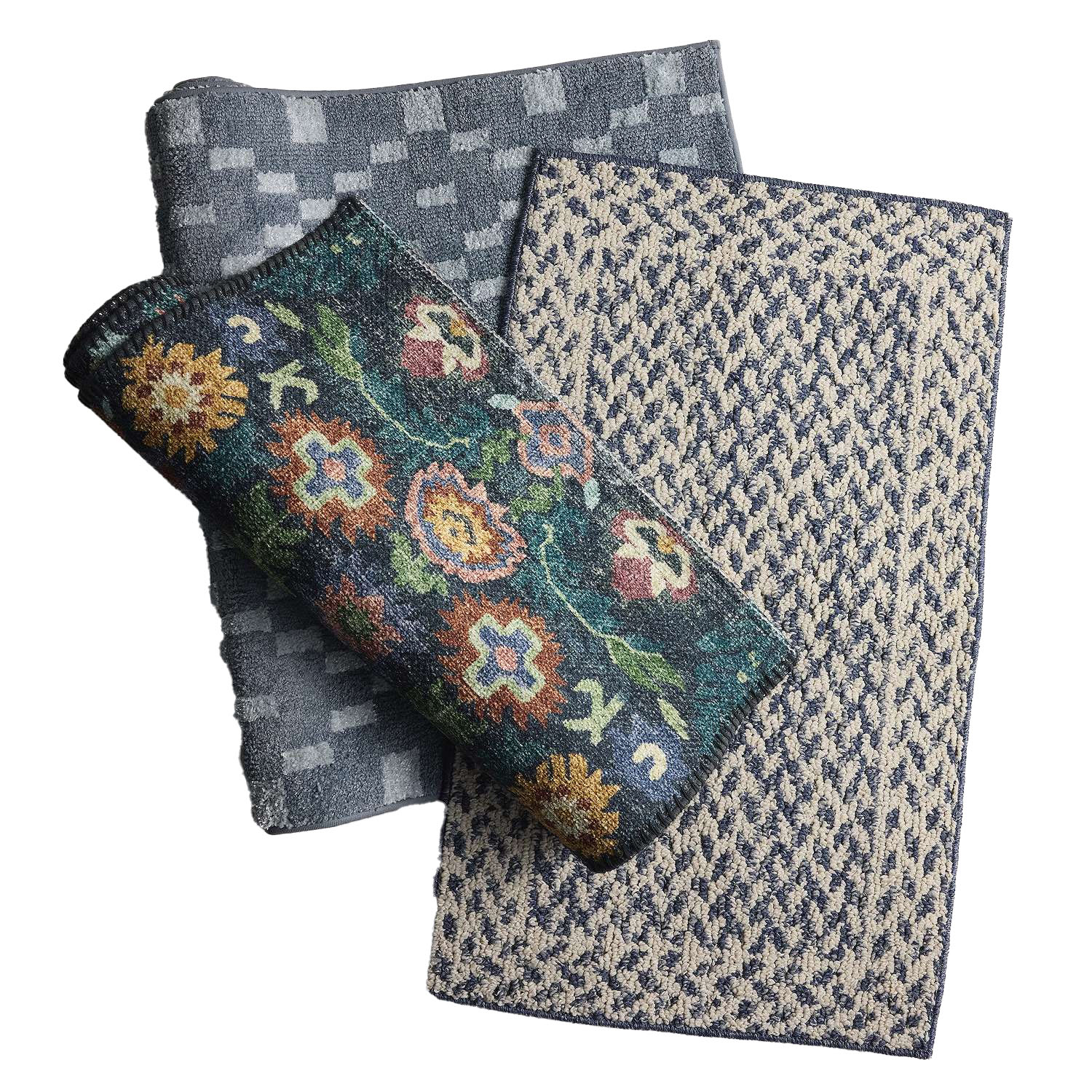 Three folded rugs with different designs: a blue geometric pattern, a beige and blue textured pattern, and a rolled-up floral pattern with multicolored flowers on a dark background.