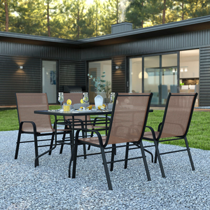 Four brown mesh stacking chairs surround a black metal table on a gravel patio, featuring glasses, a pitcher, and lemon slices. A modern black-clad house with large windows is in the background.