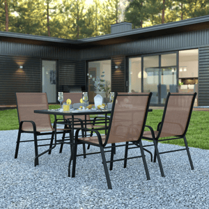 Four brown mesh stacking chairs surround a black metal table on a gravel patio, featuring glasses, a pitcher, and lemon slices. A modern black-clad house with large windows is in the background.