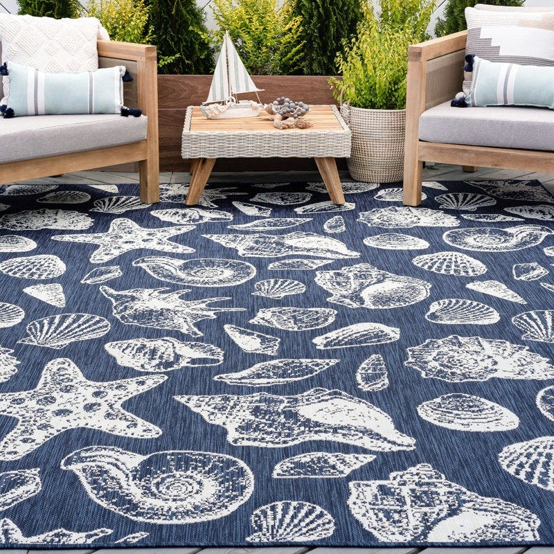 Outdoor seating area with wooden furniture and cushions, featuring a blue rug adorned with white seashell and starfish patterns; a small table with nautical decor is in the center.
