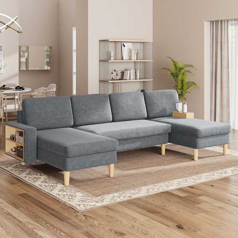 Grey L-shaped sectional sofa with wooden legs, featuring built-in storage on the armrests, set on a patterned area rug. Background includes a minimalist dining area, a bookshelf, and a potted plant.