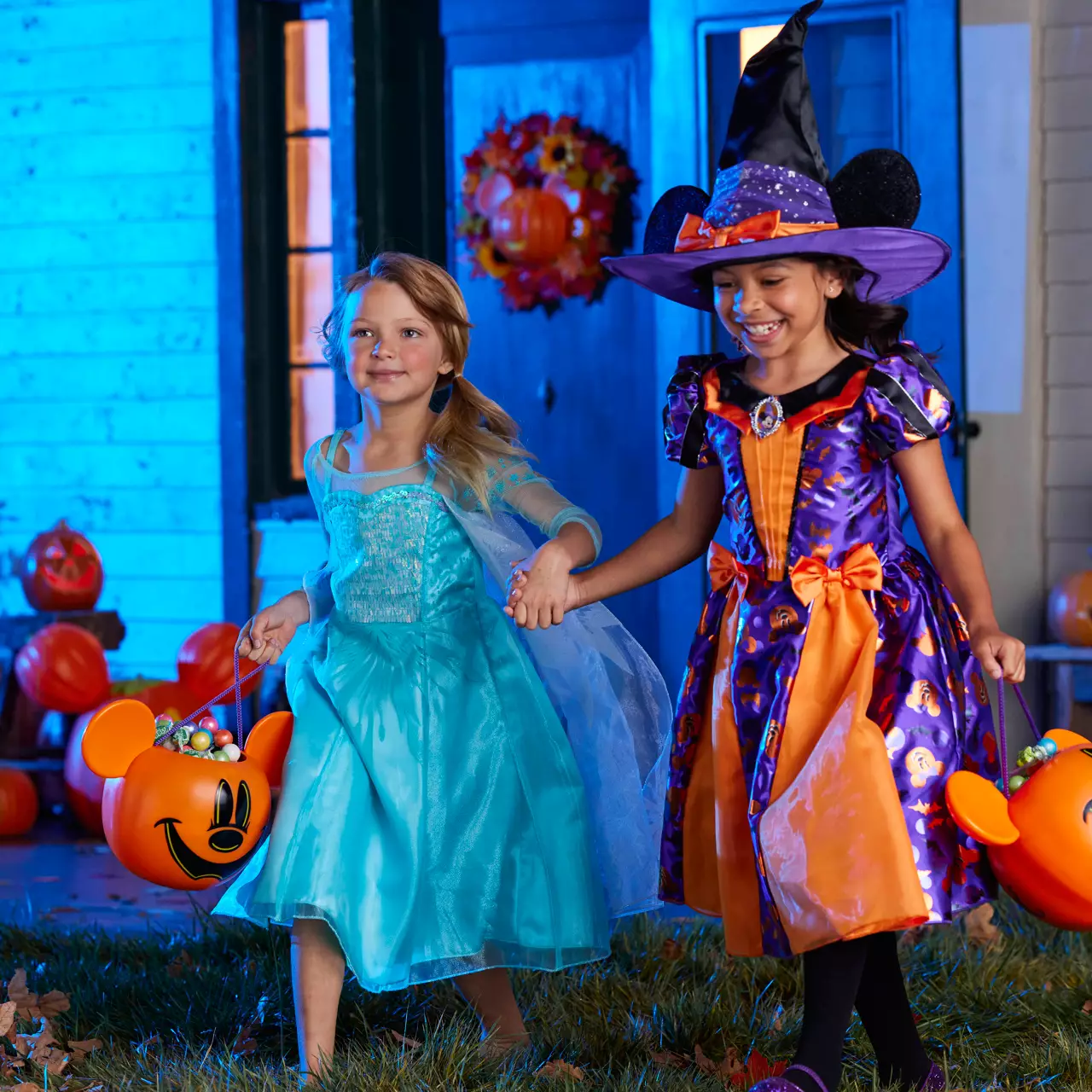A child is wearing a Minnie Mouse witch costume with a colorful dress patterned with Minnie icons, a witch's hat with mouse ears, and an orange bow. She carries a matching pumpkin-shaped treat bucket.