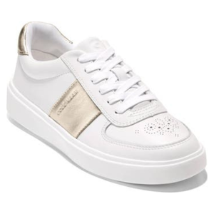 White Cole Haan Grand Crosscourt Della sneakers feature a gold metallic stripe on the side and back, with perforated detailing on the toe. They have a lace-up design and a clean, minimalist aesthetic.