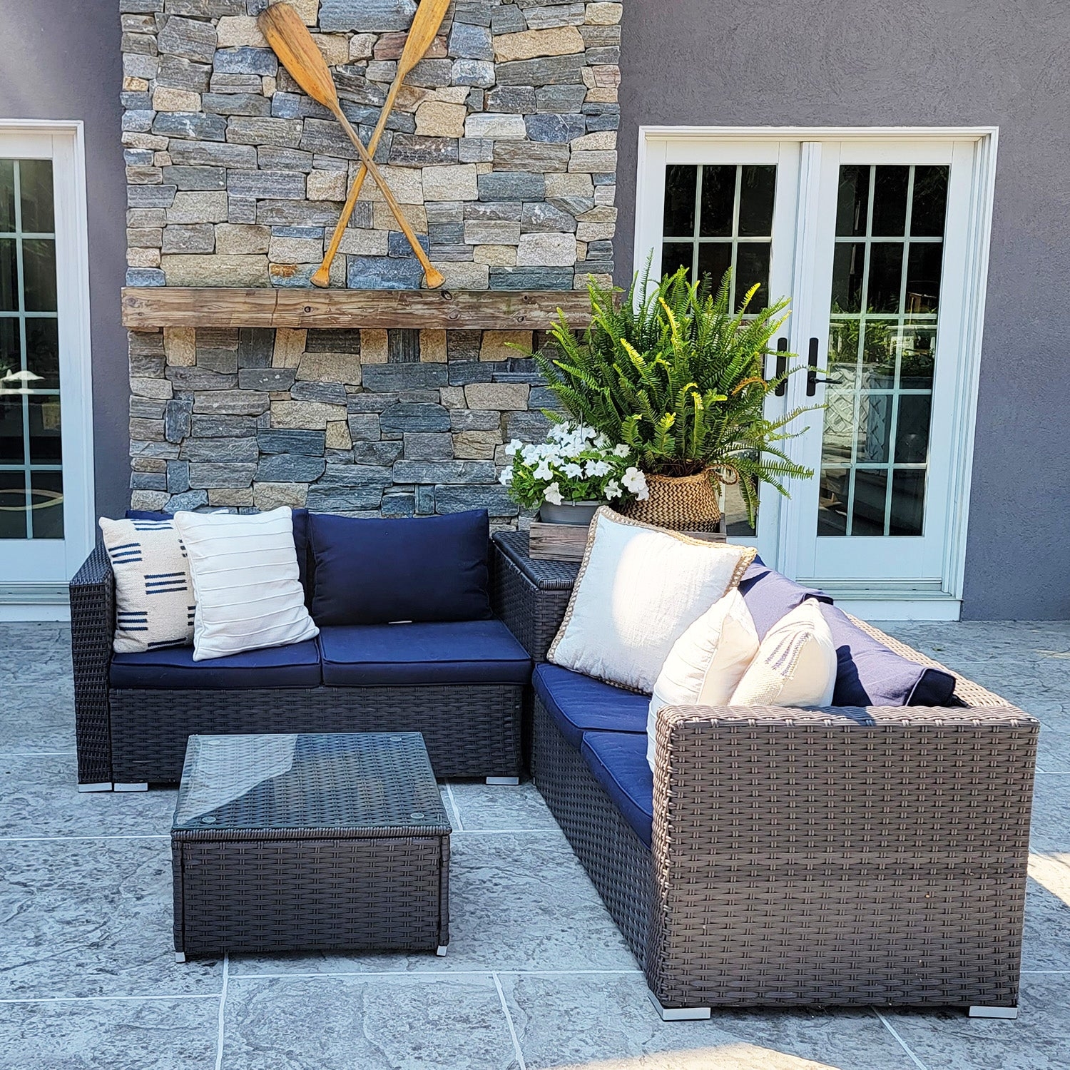 A 4-seat patio sectional set features dark wicker with navy cushions and is arranged in an L-shape. It includes a matching square glass-top coffee table and is adorned with decorative pillows.