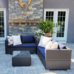 A 4-seat patio sectional set features dark wicker with navy cushions and is arranged in an L-shape. It includes a matching square glass-top coffee table and is adorned with decorative pillows.