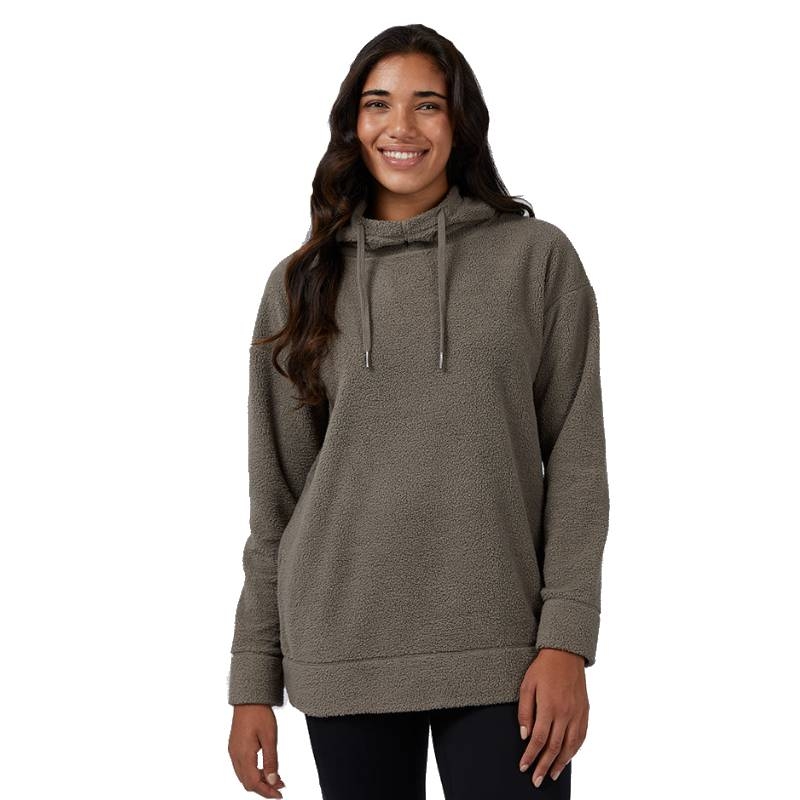 A woman is wearing a cozy, oversized sherpa hoodie in a neutral taupe color, featuring a drawstring hood and long sleeves with cuffed ends.