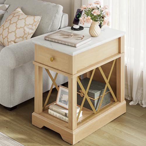 A wooden side table with a white top, featuring a drawer with a gold handle and open shelving with X-shaped accents, holds books, a framed photo, a decorative box, and a vase of flowers.