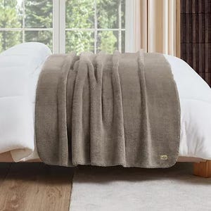A soft, taupe-colored plush throw with a cozy texture, draped over a white bed. It features a small UGG logo in the corner, indicating the brand.