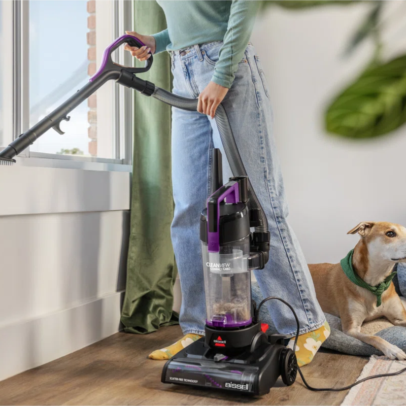 A person uses a BISSELL vacuum cleaner with a detachable hose to clean a windowsill while a dog rests on a rug nearby.