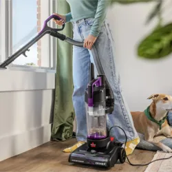 A person uses a BISSELL vacuum cleaner with a detachable hose to clean a windowsill while a dog rests on a rug nearby.
