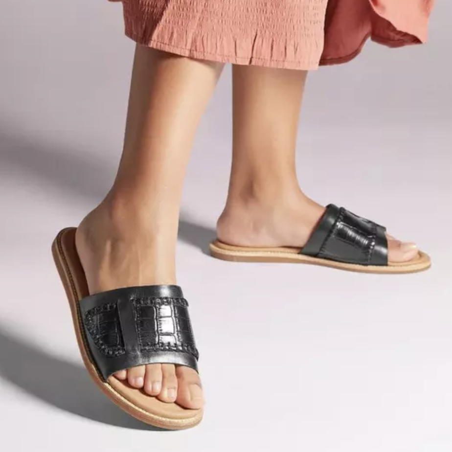 A pair of black slide sandals with a wide strap, worn with a pink skirt.
