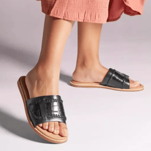 A pair of black slide sandals with a wide strap, worn with a pink skirt.