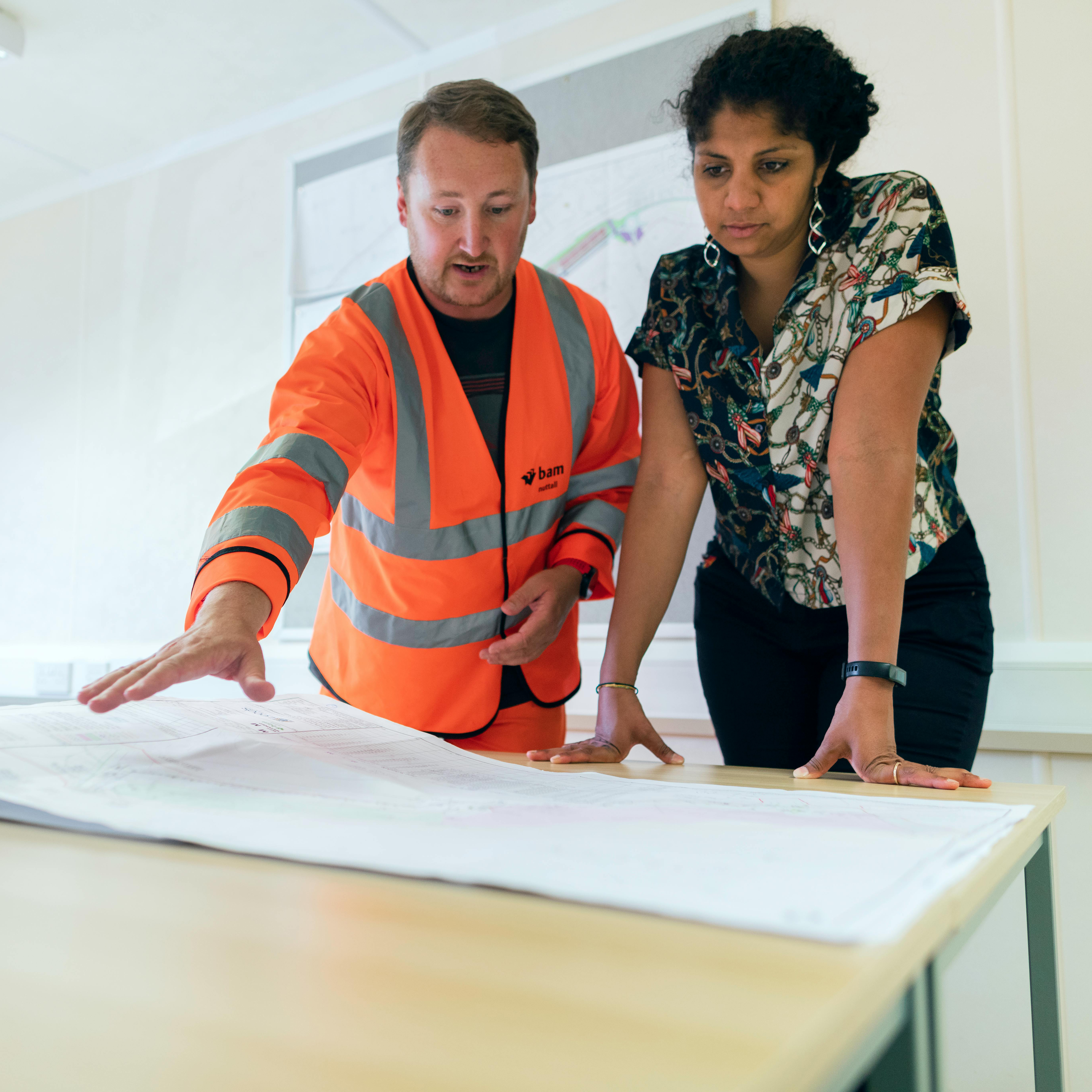 Two people, one in a hi-vis vest, are examining large blueprints on a table.