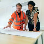 Two people, one in a hi-vis vest, are examining large blueprints on a table.