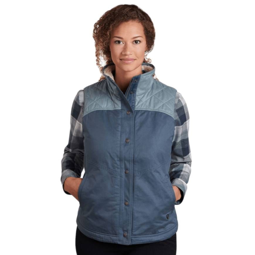 A woman models a Kuhl fleece-lined vest featuring a two-tone blue color scheme with a quilted yoke and snap-button closure. She wears a blue plaid shirt underneath the vest, which has pockets and a high collar.