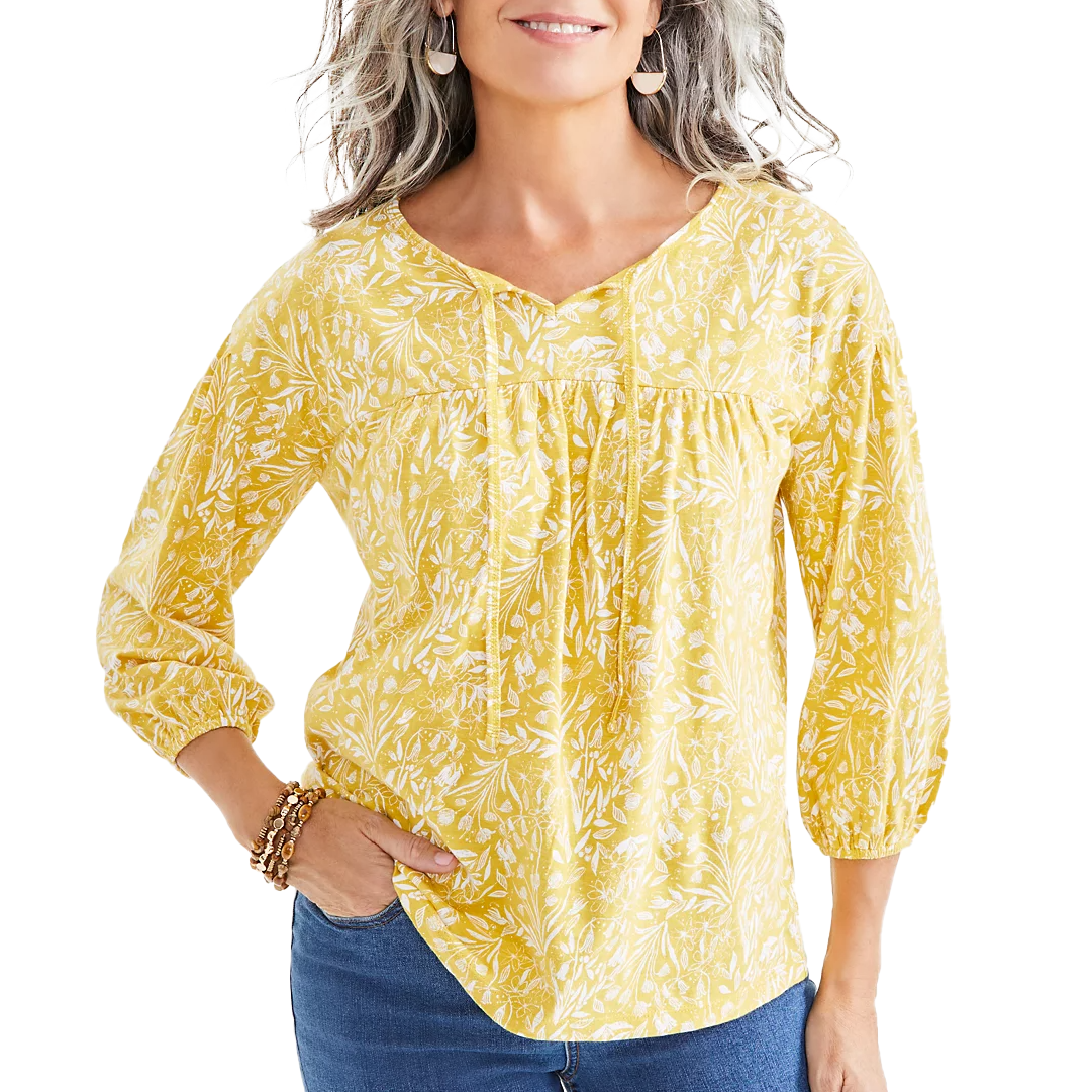 A woman is wearing a yellow floral-printed knit peasant top with three-quarter sleeves and a gathered neckline, paired with blue jeans. She accessorizes with dangling earrings and bracelet.