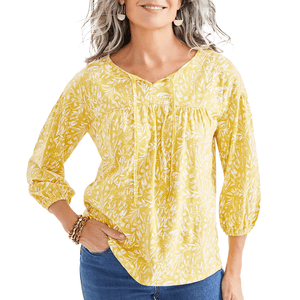 A woman is wearing a yellow floral-printed knit peasant top with three-quarter sleeves and a gathered neckline, paired with blue jeans. She accessorizes with dangling earrings and bracelet.