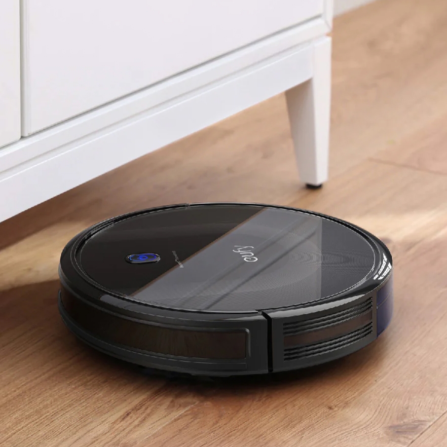 A black robotic vacuum cleaner is operating on a wooden floor near a white piece of furniture.