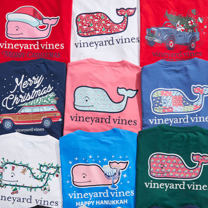 Vineyard Vines holiday-themed t-shirts featuring designs like whales in Christmas and Hanukkah motifs, decorated with festive elements such as Santa hats, menorahs, and holiday vehicles.