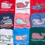 Vineyard Vines holiday-themed t-shirts featuring designs like whales in Christmas and Hanukkah motifs, decorated with festive elements such as Santa hats, menorahs, and holiday vehicles.