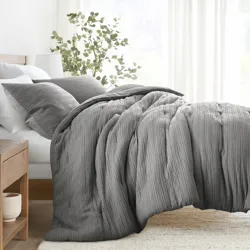 A cozy bed setup featuring a textured gray comforter and matching pillowcases, complemented by a wooden nightstand and a potted plant, in a bright and airy bedroom setting.