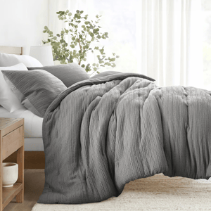 A cozy bed setup featuring a textured gray comforter and matching pillowcases, complemented by a wooden nightstand and a potted plant, in a bright and airy bedroom setting.