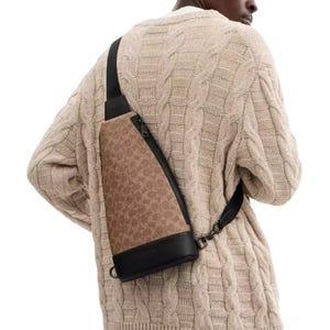 A tan and brown sling bag with a signature Coach pattern and black leather accents, worn over the shoulder. It features a zipper closure and is styled with a knitted cream sweater.