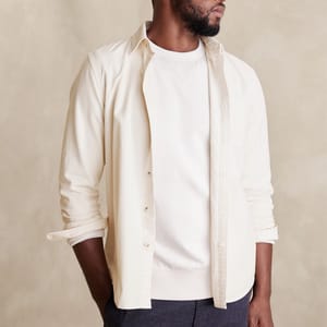 A man wearing a cream-colored button-up shirt over a white long-sleeve t-shirt paired with dark trousers.