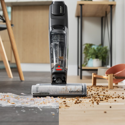 A Bissell cordless wet/dry vacuum stands upright on a wooden floor, with its motor on top and a clear dirt collection container beneath, actively cleaning scattered dry debris.