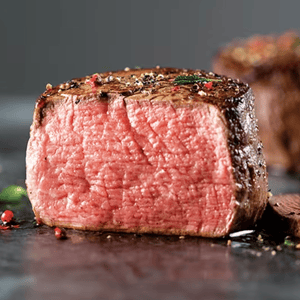 A thick cut of medium-rare steak, seasoned with coarse black pepper and garnished with herbs and peppercorns, is presented on a dark surface.