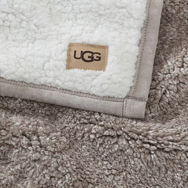 A soft, beige UGG branded blanket is shown, featuring a plush, white fleece interior and suede-like trim.