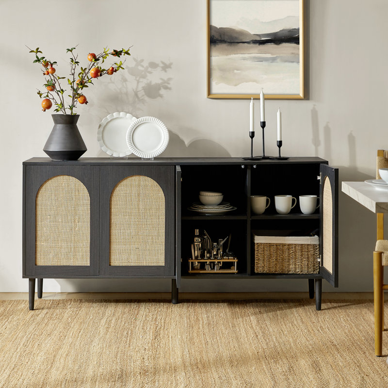 A dark wood sideboard with wicker doors stands on a woven rug, featuring open shelves displaying plates and mugs. A vase with branches, decorative plates, and candlesticks adorn the top, with a framed abstract artwork hanging above.