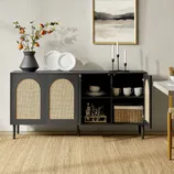 A dark wood sideboard with wicker doors stands on a woven rug, featuring open shelves displaying plates and mugs. A vase with branches, decorative plates, and candlesticks adorn the top, with a framed abstract artwork hanging above.