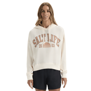 A woman models a cream-colored, waffle-knit hoodie with \“Salt Life 2003\“ printed on the front, paired with black shorts.