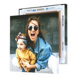 A square canvas print displaying a joyful woman in sunglasses and a child in a yellow sweater with a headband, with another canvas behind it showing similar framing.